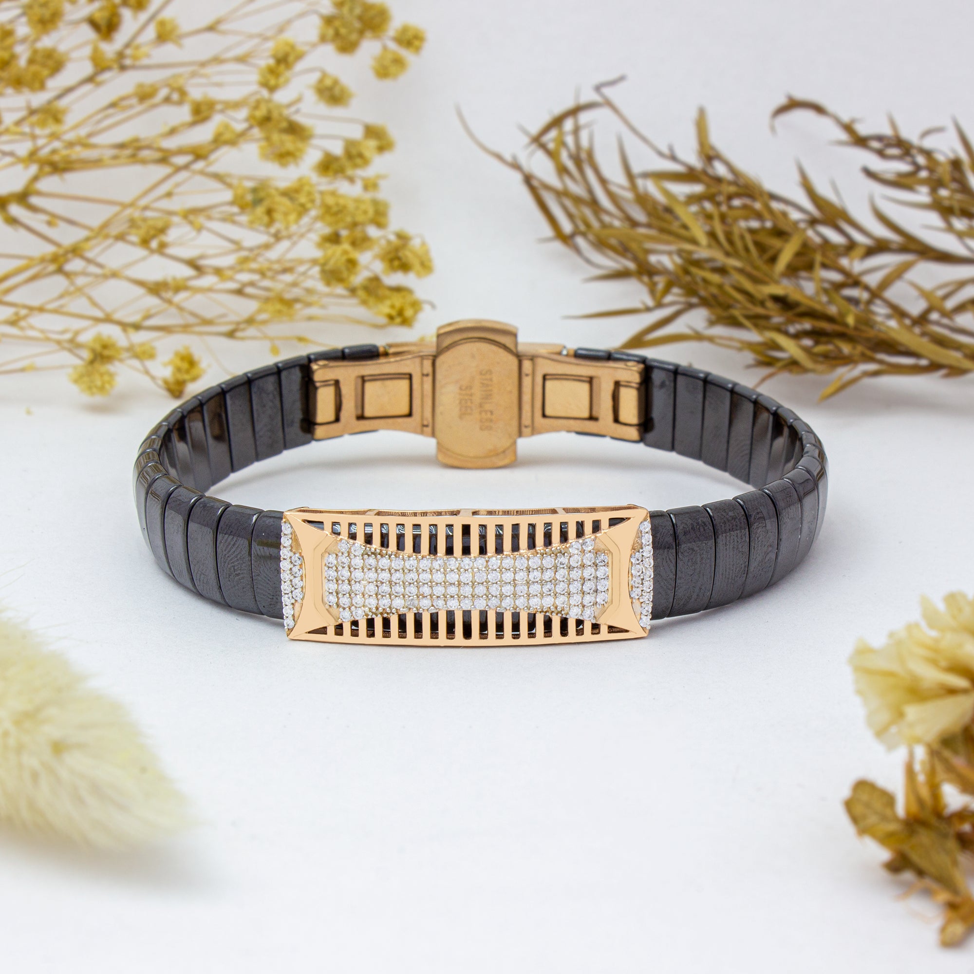 Gold and black bracelet with diamond accents on a white surface with dried flowers.